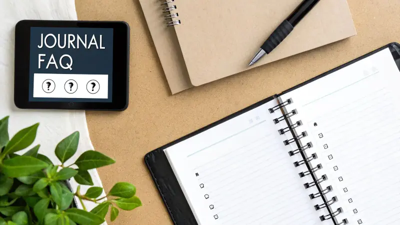 Overhead shot of a desk with a tablet displaying 'JOURNAL FAQ', notebooks, a pen, and a green plant.